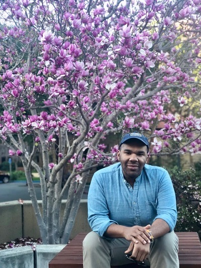 George Weddington, a fourth-year graduate student in the Department of Sociology, sits on a bench near a cherry blossom tree